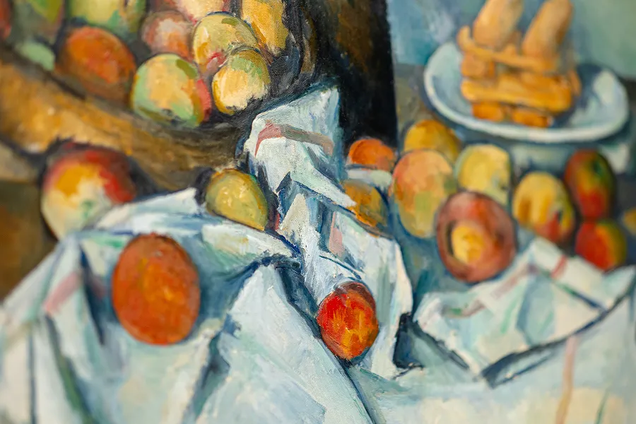 A close-up of a still life painting showing apples and oranges on a crumpled white cloth, with more fruit and a plate in the background.