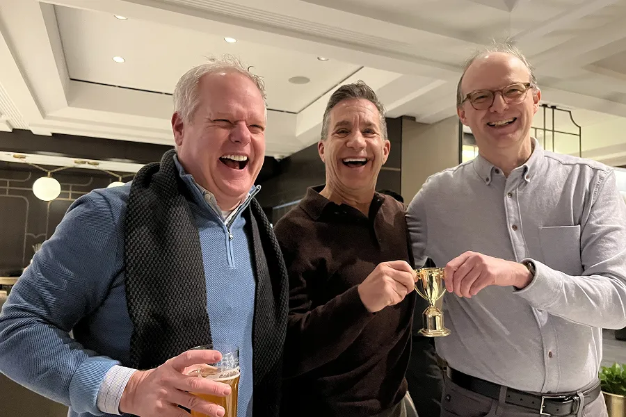 Three middle-aged men smiling, with two holding a small gold trophy and one holding a drink, standing indoors in a well-lit room.