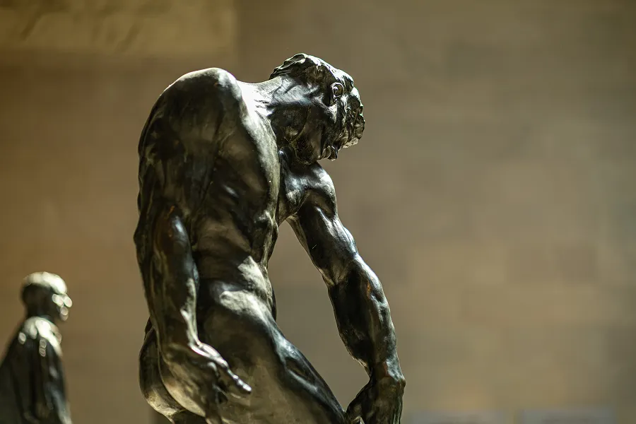 A bronze sculpture of a muscular male figure with a downward gaze and bent posture, displayed indoors with a blurred background.
