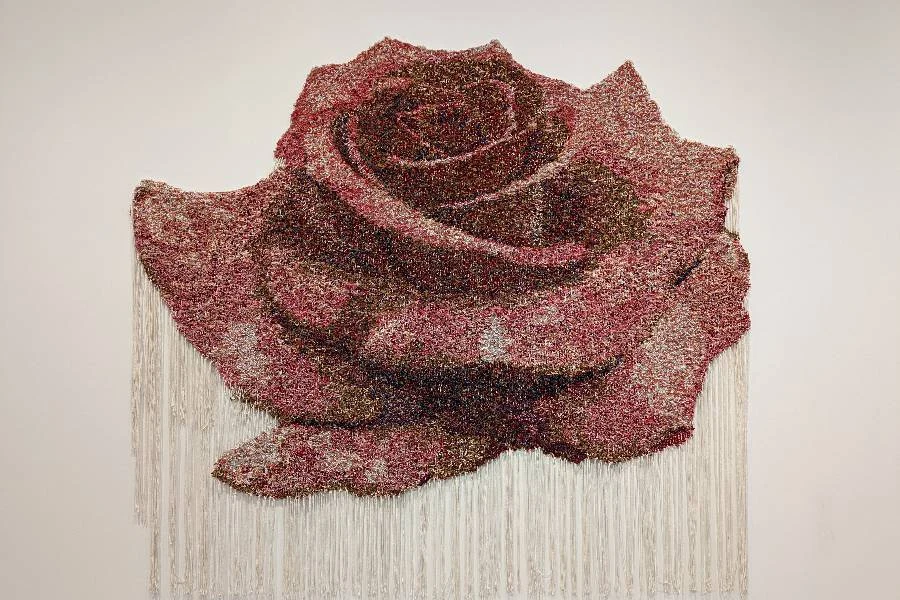 A large, textured rose artwork made of small, multicolored elements hangs on a wall, with white fringe strands hanging from the bottom edge.