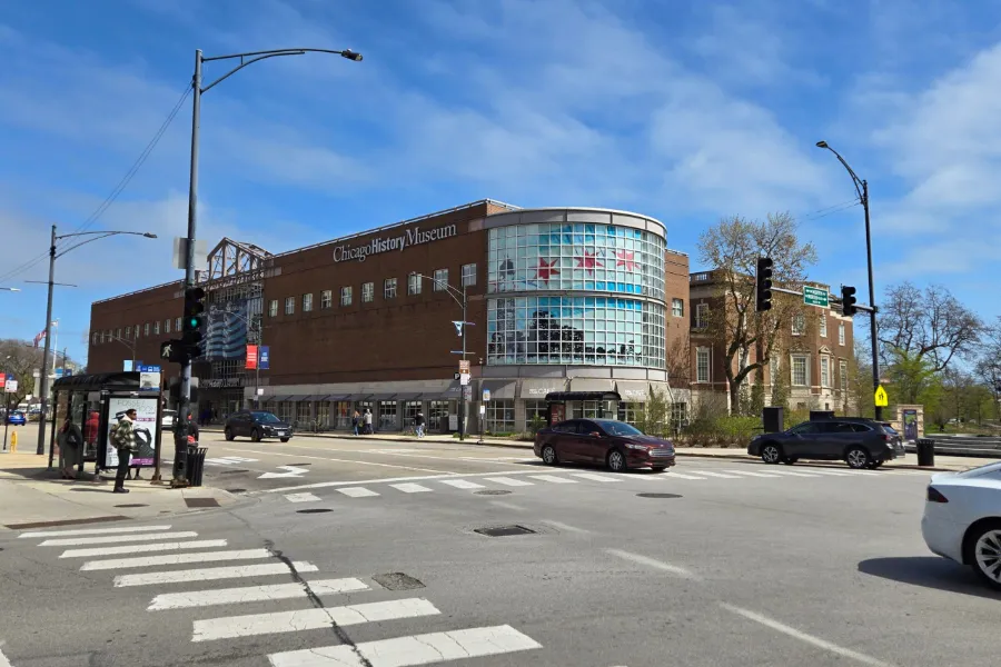 Chicago History Museum building at a street corner with cars, pedestrians, and a blue sky in the background.